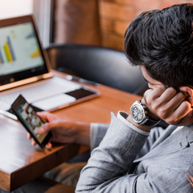 man sat a desk browsing his phone, while on the desk his computer is on.