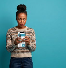 An image of a young woman with her dark hair in a bun against a bright blue background. She is wearing a patterned beige sweater and blue jeans. She has a concerned and confused expression on her face as she looks down at a large smartphone she is holding with both hands. The phone screen displays a webpage with text and images.