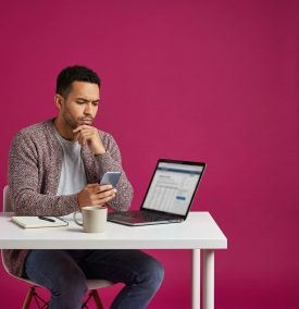 A young man sits at a white desk against a vibrant magenta background, appearing deep in thought. He is wearing a mauve textured cardigan over a grey t-shirt and blue jeans. He holds a smartphone in one hand while resting his other hand on his chin, looking toward an open laptop on the desk. Next to the laptop are a white coffee mug and a spiral notebook with a pen.