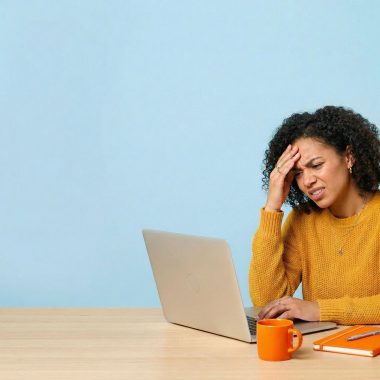 A woman with curly hair expresses stress or a headache while looking at a laptop screen showing design software. She is seated at a wooden desk against a plain blue background, with an orange mug and notebook nearby.