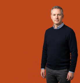 A man with short grey hair wearing a navy blue sweater over a light blue shirt, standing confidently against a solid orange background.