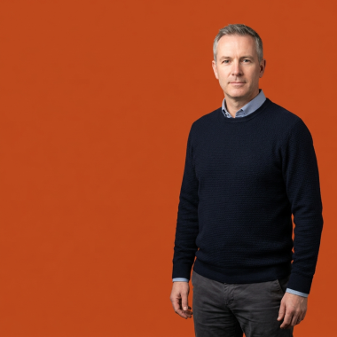 A man with short grey hair wearing a navy blue sweater over a light blue shirt, standing confidently against a solid orange background.