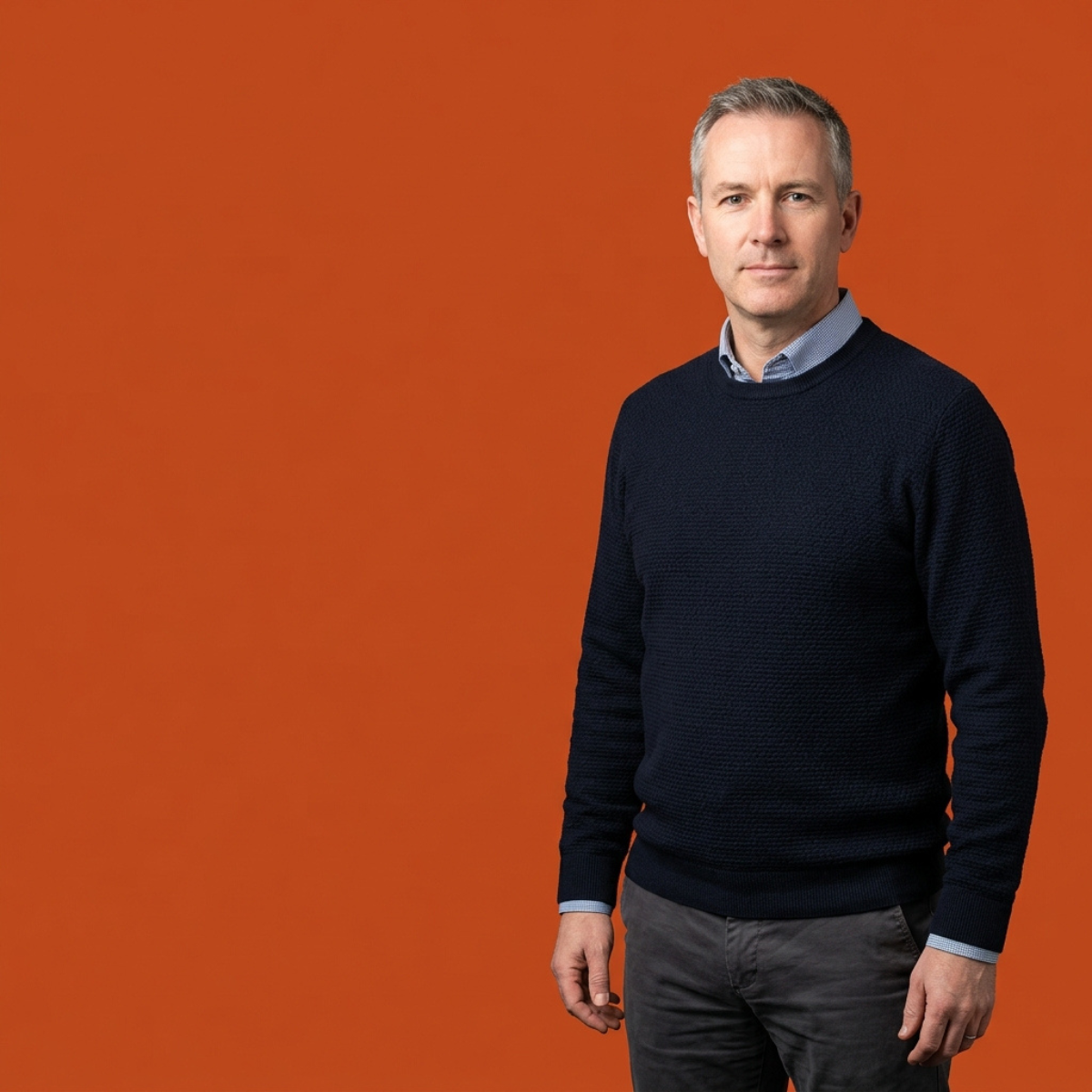 A man with short grey hair wearing a navy blue sweater over a light blue shirt, standing confidently against a solid orange background.