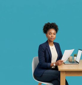 A professional woman in a navy blazer sitting at a wooden desk, holding a document and looking at the camera with a laptop and coffee mug in a minimalist office setting.