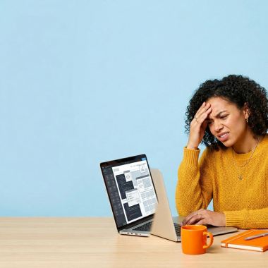 A woman with curly hair expresses stress or a headache while looking at a laptop screen showing design software. She is seated at a wooden desk against a plain blue background, with an orange mug and notebook nearby.