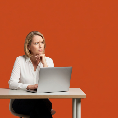 A woman with blonde hair sitting at a desk with a laptop, looking thoughtful or concerned against a solid orange background.