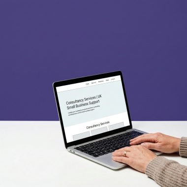 A close-up shot of a person's hands typing on a silver laptop against a dual-tone background of white and deep purple. The laptop screen displays a website titled "Consultancy Services | UK Small Business Support." The person is wearing a textured beige sweater and a simple gold wedding band on their ring finger.