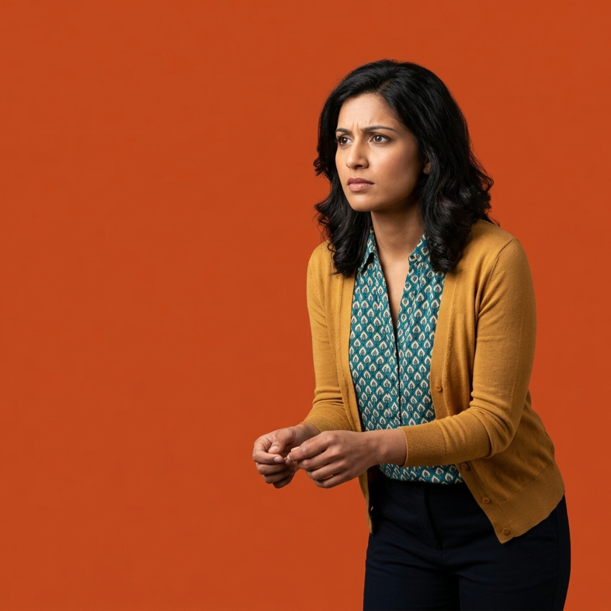 A woman with dark wavy hair looking concerned and leaning forward against a solid orange background, wearing a yellow cardigan over a patterned shirt.