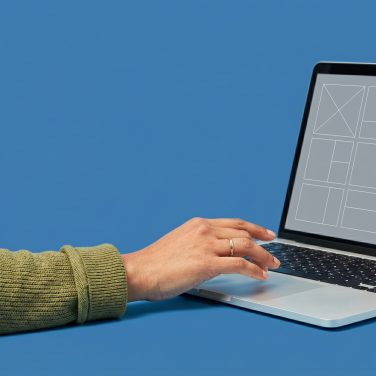 Close-up of a hand near a laptop keyboard, casual sleeve, plain medium blue background.