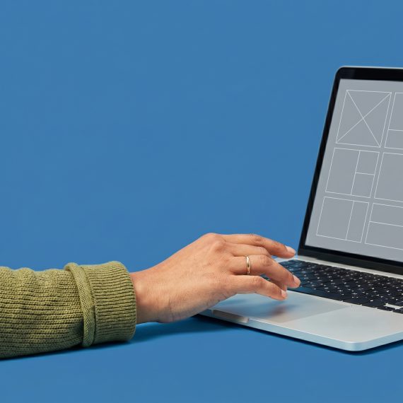 Close-up of a hand near a laptop keyboard, casual sleeve, plain medium blue background.