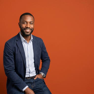 A smiling, confident Black man in his 40s wearing a navy blazer and striped shirt, posed against a solid burnt orange background with significant negative space.