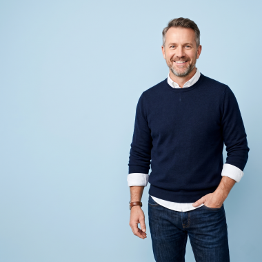 A smiling middle-aged man with grey hair and beard, wearing a navy blue sweater and jeans, stands against a light blue background.
